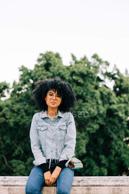 Jeune Afro-Américaine à la mode en jeans et sur le toit d'une récolte assise sur un parapet de pierre et regardant la caméra — Photo de stock