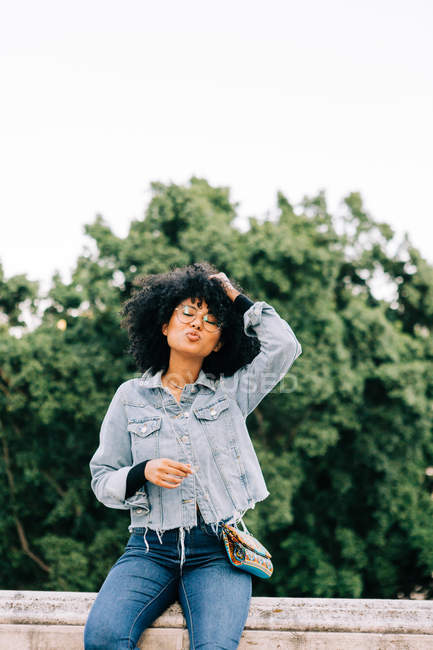 Junge trendige Afroamerikanerin in Jeans und bauchfreiem Top sitzt auf Steinbrüstung — Stockfoto