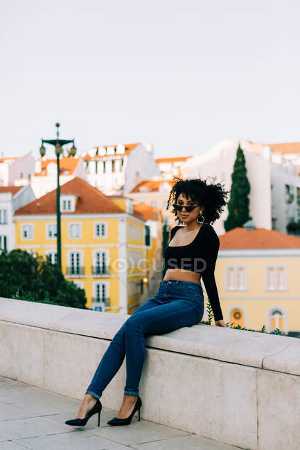 Молода афроамериканська жінка в джинсах і урожаї зверху сидить на кам'яному парапеті і дивиться за сонцезахисними окулярами. — стокове фото