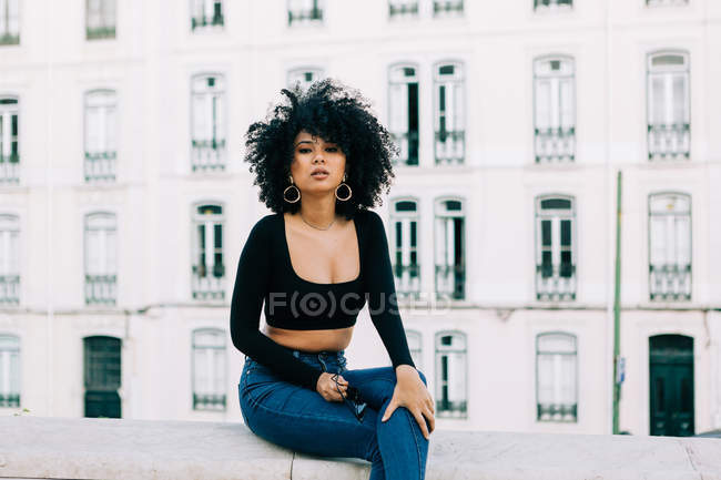 Молодая трепетная афроамериканка в джинсах и кроссовках сидит на каменном парапете и смотрит в камеру — стоковое фото