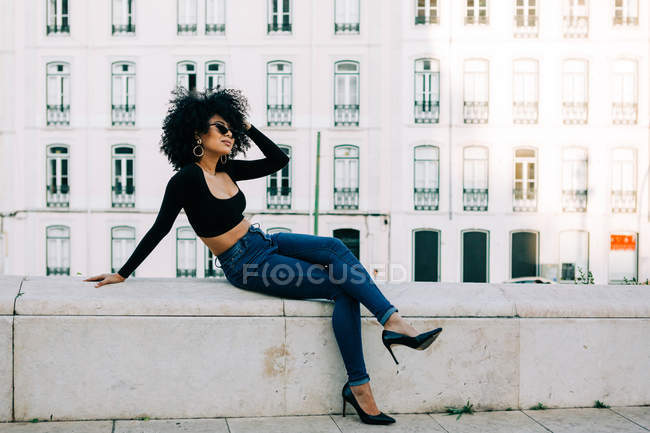 Jeune Afro-Américaine à la mode en jeans et sur le toit d'une récolte assise sur un parapet de pierre et regardant par-dessus des lunettes de soleil — Photo de stock