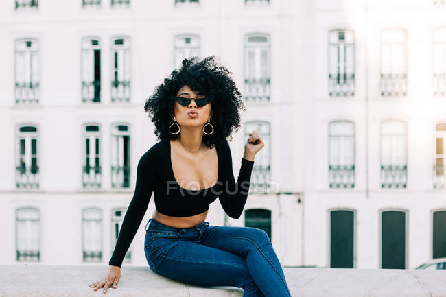 Молодая трепетная афроамериканка в джинсах и кроссовках сидит на каменном парапете и смотрит на солнцезащитные очки — стоковое фото