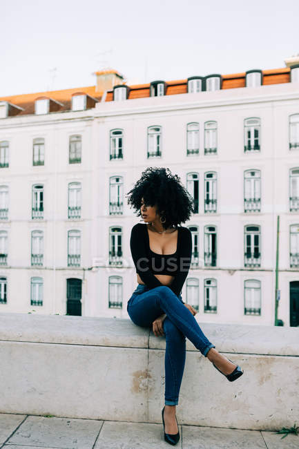 Young trendy African American woman in jeans and crop top sitting on stone parapet — Stock Photo