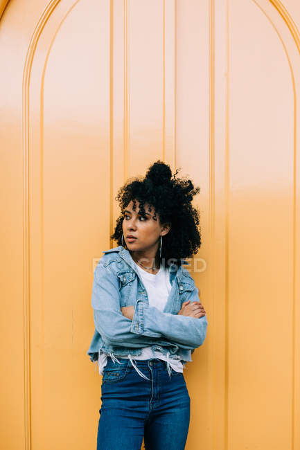 Jovem afro-americana em jeans e jaqueta de ganga apoiada na porta amarela e olhando para longe — Fotografia de Stock