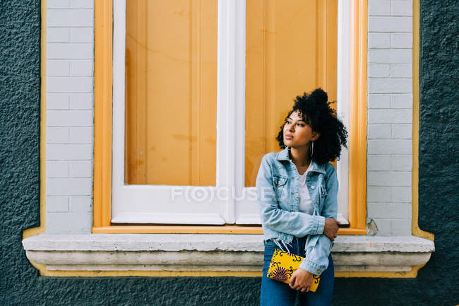 Mulher étnica na moda em jeans e jaqueta jeans segurando bolsa de moda e inclinando-se no peitoril da janela brilhante ao ar livre — Fotografia de Stock