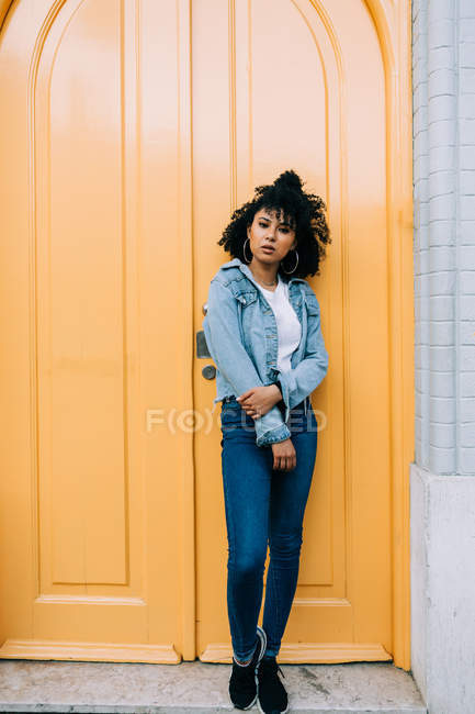 Joven mujer afroamericana en jeans y chaqueta de mezclilla apoyada en la puerta amarilla y mirando a la cámara - foto de stock