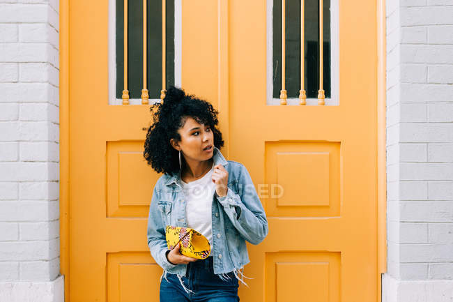 Young African American woman in jeans and denim jacket leaning on yellow door, holding clutch and looking away — Stock Photo