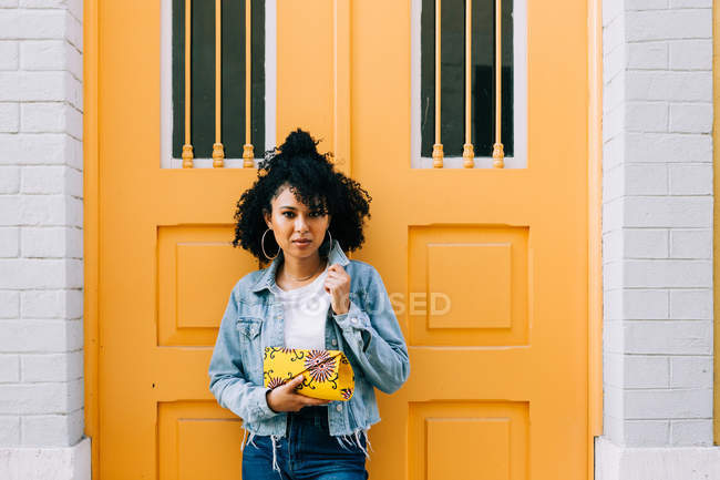 Giovane donna afroamericana in jeans e giacca di denim appoggiata alla porta gialla, che tiene la pochette e guarda la macchina fotografica — Foto stock