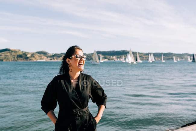 Vista lateral de la joven atractiva mujer en traje negro de pie por mar en Lisboa - foto de stock