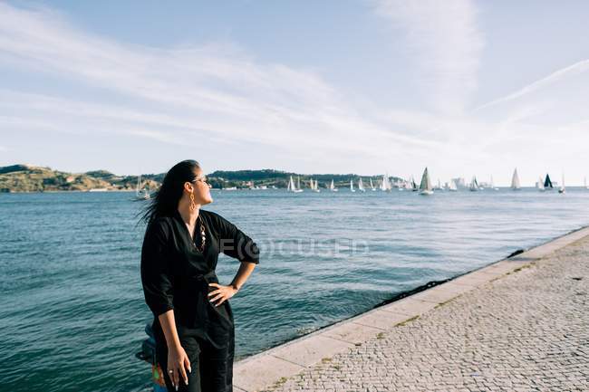 Seitenansicht einer jungen attraktiven Frau im schwarzen Outfit, die am Meer steht und sich auf eine Stange in Lissabon stützt — Stockfoto