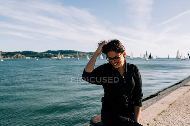 Vista lateral de la joven atractiva mujer en traje negro de pie junto al mar y apoyado en el poste en Lisboa - foto de stock