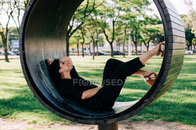 Vista lateral de la mujer acostada en un banco creativo en forma de anillo y sonriente en Lisboa en verano - foto de stock