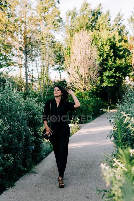 Модная молодая женщина в черном костюме идет по асфальтированной тропинке и наслаждается летним днем в Лиссабоне — стоковое фото