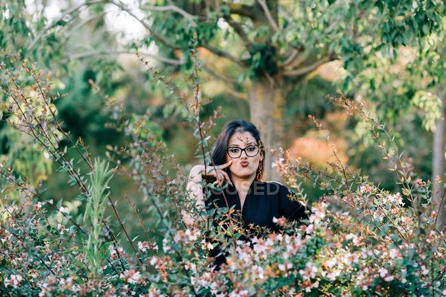 Mulher morena bonita em óculos de pé entre arbustos florescentes no parque e olhando para a câmera em Lisboa — Fotografia de Stock