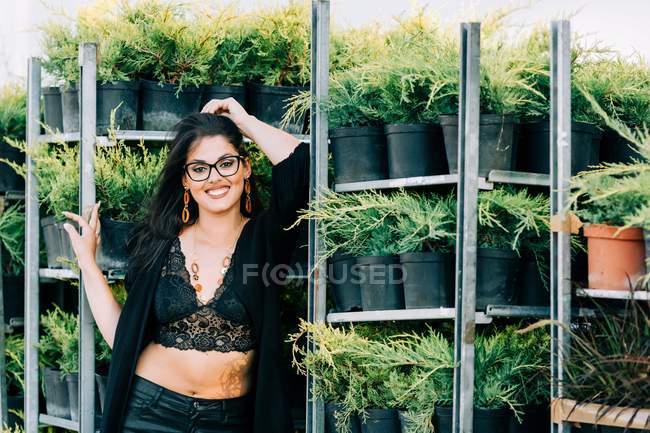 Donna alla moda in pizzo crop top appoggiato su rack con piante in vaso e guardando la fotocamera a Lisbona — Foto stock