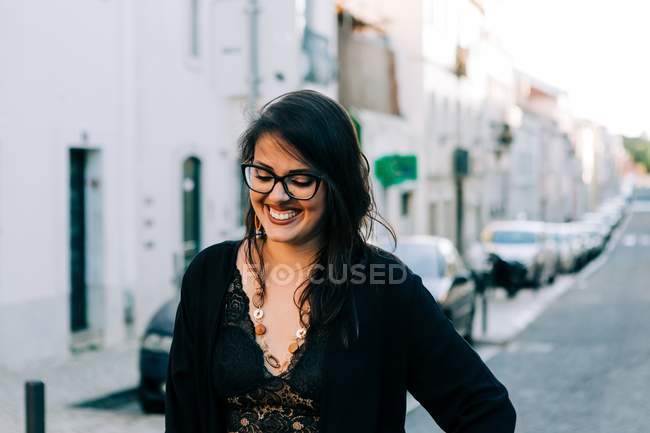 Mujer joven de moda en traje negro de pie en el paso de peatones con las manos en las caderas en Lisboa y sonriendo - foto de stock