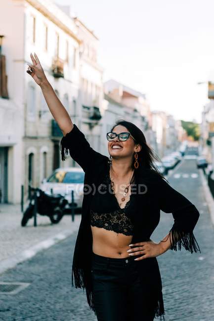 Giovane donna alla moda in abito nero in piedi sul marciapiede con le mani sui fianchi a Lisbona e sorridente — Foto stock