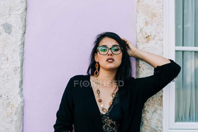 Atractiva joven mujer en gafas y blusa negra de pie junto a la colorida pared vintage y mirando a la cámara en Lisboa - foto de stock
