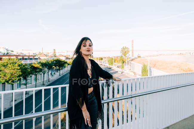 Стильная женщина в черном наряде стоит у моста с городским пейзажем в Лисбоне в солнечный день — стоковое фото