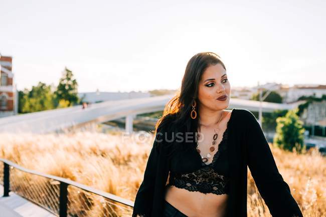 Beautiful stylish woman in black outfit standing by bridge with city landscape in Lisbon on sunny day — Stock Photo