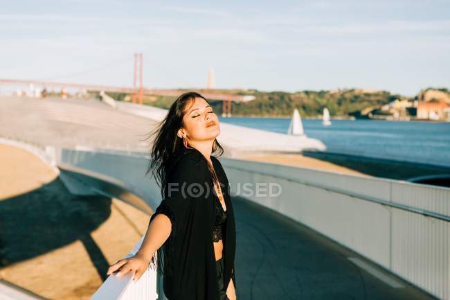Schöne stilvolle Frau im schwarzen Outfit steht an einem sonnigen Tag an einer Brücke mit Stadtlandschaft in Lissabon — Stockfoto