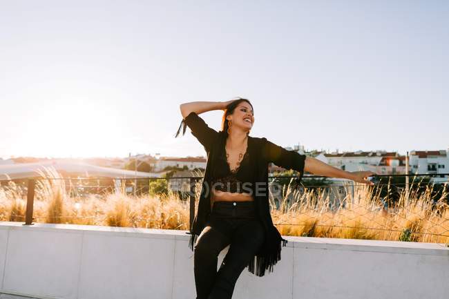 Beautiful stylish woman in black outfit sitting by bridge with city landscape in Lisbon on sunny day — Stock Photo