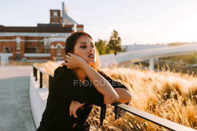 Beautiful stylish woman in black outfit standing by bridge with city landscape in Lisbon on sunny day — Stock Photo