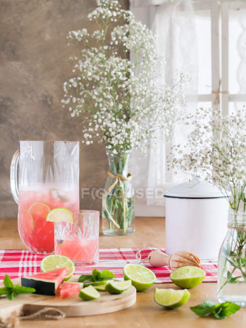 Cold jug of iced pink lemonade with watermelon and limes on rustic kitchen table next to bunch of gypsophila flowers in daylight — Stock Photo