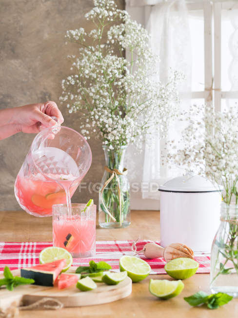 Persona vertiendo limonada de sandía fría en el vidrio del frasco en la mesa de la cocina rústica al lado del ramo de flores de gypsophila - foto de stock