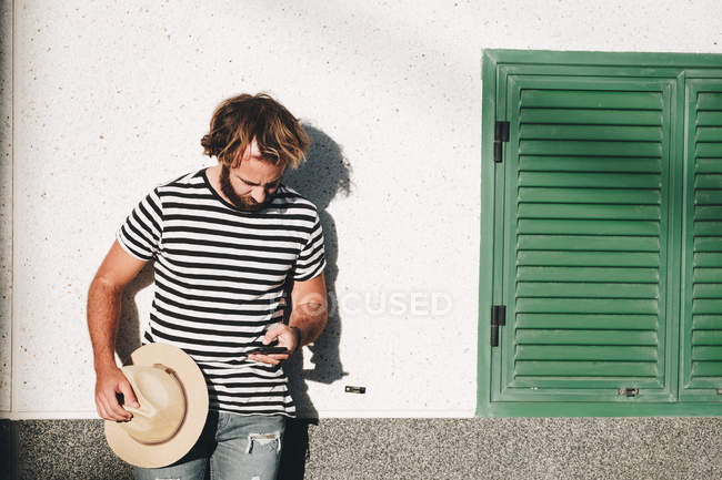 Bearded man in striped t-shirt standing by wall, holding hat and using mobile phone in La Restinga, El Hierro — Stock Photo