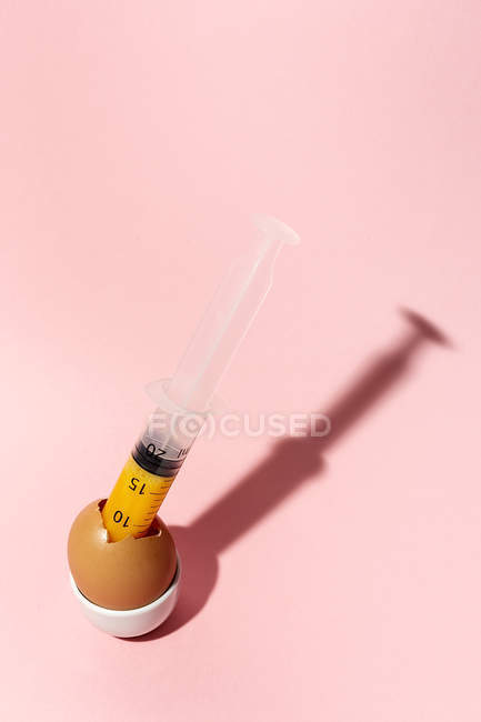 Huevo de cocina en taza de huevo con jeringa sacando yema cruda amarilla sobre fondo rosa - foto de stock