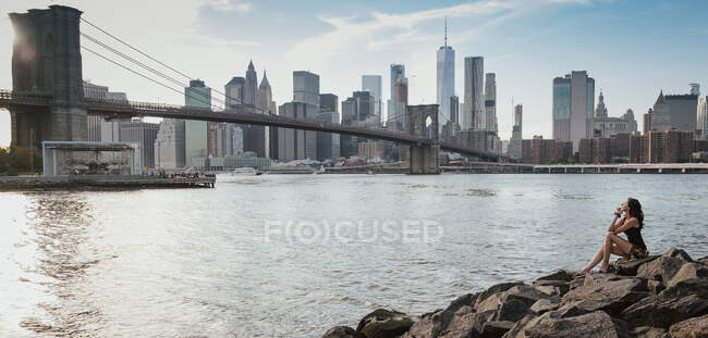 Donna seduta su rocce a Brooklyn guardando il ponte e Manhattan in una giornata di sole — Foto stock