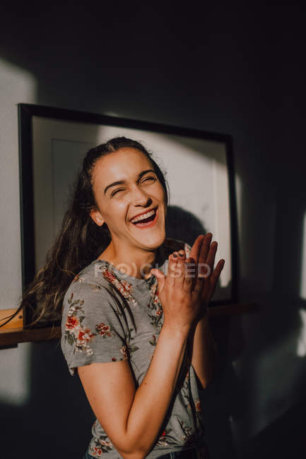 Aufgeregte junge Frau im T-Shirt lacht und klatscht in die Hände, während sie neben der weißen Wand steht — Stockfoto