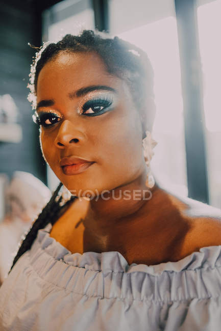 Portrait de belle jeune femme noire courbée avec un maquillage lumineux en robe hors épaule debout dans un café au coucher du soleil — Photo de stock
