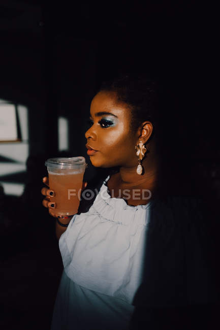 Portrait de vue latérale de belle jeune femme noire incurvée avec un maquillage lumineux en robe hors épaule ayant une boisson froide — Photo de stock