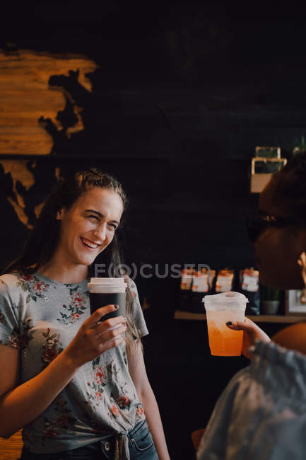 Вид збоку веселі багаторасові молоді випадкові жінки сміються і п'ють каву, сидячи біля вікна в кафе на заході сонця — стокове фото