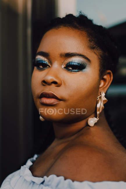 Attraktive schwarze junge Frau mit hellem Make-up im schulterfreien Kleid, die bei Sonnenuntergang auf der Straße steht und in die Kamera blickt — Stockfoto