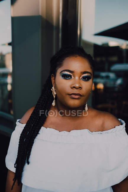 Attractive black young woman with bright makeup in off-shoulder dress standing on street on sunset, looking at camera — Stock Photo
