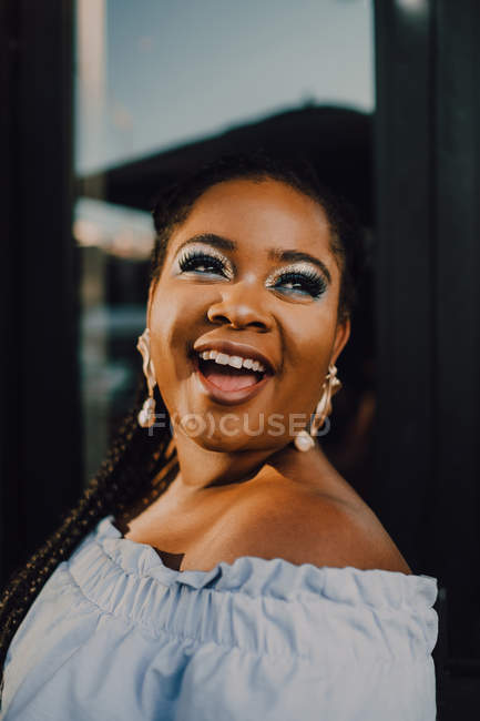 Attractive black young woman with bright makeup in off-shoulder dress standing on street on sunset, looking away — Stock Photo