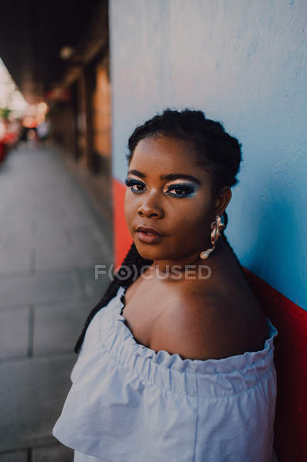 Attrayant moderne jeune femme noire jeune avec un maquillage lumineux en robe hors épaule regardant la caméra — Photo de stock
