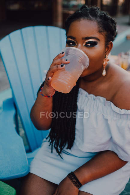 Жизнерадостная африканская молодая женщина, пьющая холодный напиток, сидя в хипстерском кафе, отворачиваясь — стоковое фото