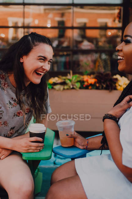 Alegre multirracial jóvenes mujeres casuales hablando y bebiendo café mientras se sienta junto a vibrante mesa en hipster café - foto de stock