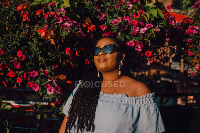 Привлекательная черная стильная молодая женщина в платье без плеча и солнечных очках, наслаждающаяся солнцем, стоя на улице — стоковое фото