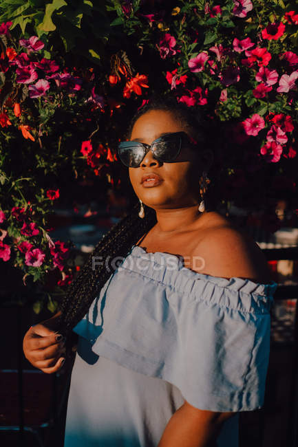 Attractive black stylish young woman in off-shoulder dress and sunglasses enjoying sunshine while standing on street — Stock Photo