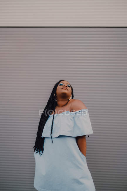 Jolie jeune femme noire avec un maquillage lumineux en robe hors épaule debout sur un mur vide, regardant vers le haut — Photo de stock