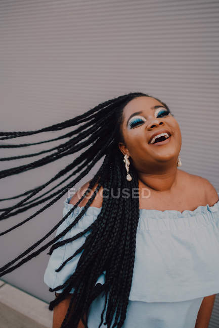 Smiling black young woman with bright make-up in off-shoulder dress flipping braids while standing on street — Stock Photo