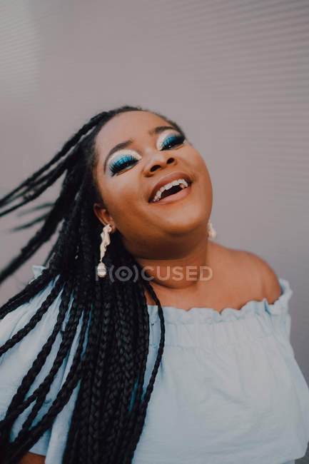Smiling black young woman with bright make-up in off-shoulder dress flipping braids while standing on street — Stock Photo