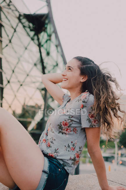 Calm content casual young woman in shorts and t-shirt enjoying sunshine while sitting on concrete parapet, looking away — Stock Photo