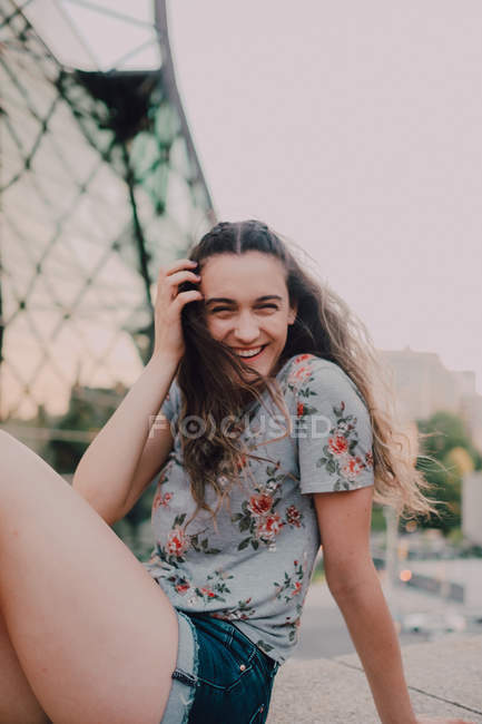 Спокойная девушка в шортах и футболке, наслаждаясь солнцем, сидя на бетонном парапете, глядя в камеру — стоковое фото