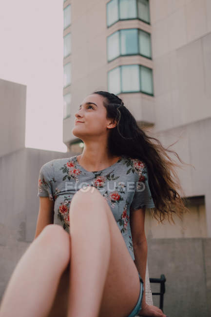 Calmo conteúdo casual jovem mulher em shorts e t-shirt desfrutar de sol enquanto sentado no parapeito de concreto, olhando para longe — Fotografia de Stock